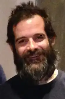 A middle-aged man with brown hair and a thick brown beard smiling at the camera.