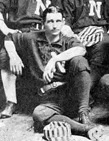 A black and white photograph of a man sitting on the floor wearing a dark baseball uniform with a white "N" on the chest. A striped cap rests at his feet.