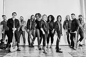 A group of eleven musicians standing on a laminate hardwood floor before a large white backdrop with a window to the viewer's right side. Some are holding instruments such as trumpets, saxophones, electric guitars an drum sticks, while others are simply present.