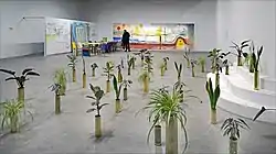 Depicts an installation by Sammy Baloji containing multiple plants in shiny containers spread out across the floor.  The background piece an installation by Fabrice Hyber.