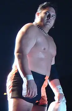 An adult Samoan male posing while wearing orange and black wrestling trunks.