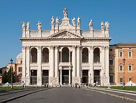 Ornate facade of the basilica at night with columns, main door, and statues of the twelve Apostles on the roofline, with a Latin inscription below them