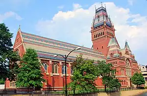 A ground-level exterior view of a large, highly ornate 19th-century building. Its main body is longer than it is high, with a single very tall story of red-orange brick with tall stained-glass windows. The steep and tall slate roof is patterned in pale shades of blue, light brown, and red-orange, arranged in broad horizontal stripes of widely varying heights. A tower rises to the right of the building's center.