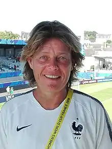 Headshot of a grey-haired women with a yellow sash, with a football ground in the background