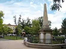 a photograph of several people sitting on a banco surrounding a stone obelisk, with pigeons all around