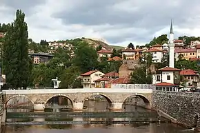 The Babića bašča local community, with Šeher-Ćehaja Bridge in front, Inat kuća just behind it, in front of the minaret which on the other hand belongs to the Vekil-Harač mosque. Cemetery Alifakovac can be seen on the right side in the background, while the fortress on the hill in background, left on the photo, is the Bijela Tabija.