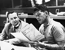 Two men seated at a desk, both reading from a large book the younger man is holding