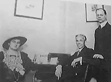 A photograph of a seated woman and man in front of a desk with a standing man behind them and to the right.