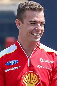 A man in his mid-twenties wearing a red zip T-shirt with a white colour scheme on the shoulders with sponsors logos and looking away from the camera with a smile