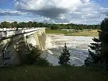 Dam wall shown with water flowing out three of the five floodgates. Spectators are gathered on the viewing area on the top of the wall watching the water flow past.