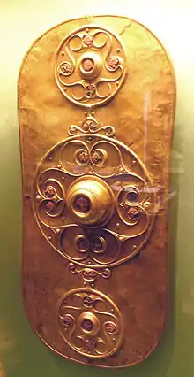 Golden shield dominated by three circular decorations. The larger central decoration has a large boss surrounded by a curlicue pattern. The smaller decorations above and below it are similar.