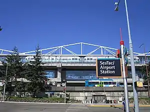 Elevated train station seen above a freeway on an embankment. A bus stop can be seen below the embankment and across a road from a sign reading "SeaTac/Airport Station" in the foreground.