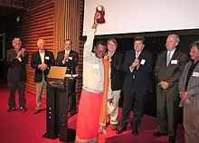 A man from Madagascar holds a trophy above his head, while people clap behind him.