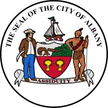 A black circle with white fill is shown with the coat of arms at center. Above the coat of arms, following an arced path, are the letters "The Seal of the City of Albany"