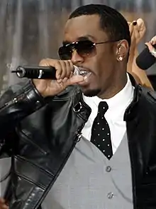 An African-American male dressed in sunglasses, a white dress shirt, grey vest, black tie and black jacket speaks into a microphone