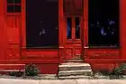  oil painting of a 200-year-old building with 2 large pane-glass windows and 2 doors, steps, weeds—painted deep red with black windows mirroring blue sky