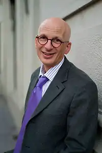 Picture of a white, bald-headed person in his 50s with brown-eyes (looking directly forward) and wearing a black shirt. The person's face is taken behind a red-orange background.