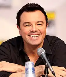 A man with short, black hair, wearing a black shirt, with slight stubble, leaning forward slightly while speaking into a microphone.