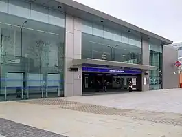 A grey-tiled building with a rectangular, dark blue sign reading "SHEPHERD'S BUSH" in white letters all under a light blue sky