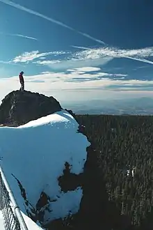 A hiker stands on a rock mound with snow surrounding him; forest can be seen below the mountain