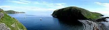 Two yachts lie in a dark blue cove set amongst green cliffs and a stony shore.