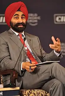 Shivinder Mohan Singh wearing a grey business suit, white shirt, red tie and turban, sitting in a chair with microphone to the right, looking at camera and appearing to speak to audience