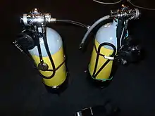 A pair of cylinders showing the regulators set up for sidemount diving. Each regulator has a short low-pressure inflator hose projecting towards where the diver's body would be, and the DV hoses are stowed under bungees. The submersible pressure gauges are on short hoses aligned with the cylinder axes.