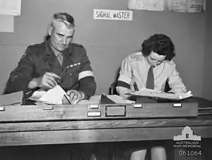 Two soldiers, one male, one female, sit side by side doing paper work at a desk. They are wearing brassards and the sign behind them says: "signal master".