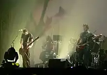 All-male group on stage, containing two MCs, two keyboardists, and a guitarist. Some of the crowd with hands in the air present in the foreground