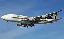 A large mostly-white four-engine jet airliner with golden stylized bird design, on approach towards left of screen with landing gear extended