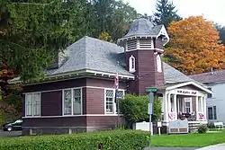 A brown wooden building with a gray peaked roof and tower and porch in front. Behind it are trees. One on the right is bright orange with autumn color