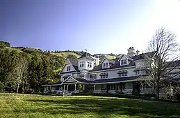 A large white house with black roofing in front of green hills and forests.