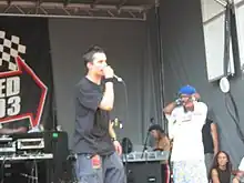 Knee high portrait of male in his twenties, facing right wearing a black t-shirt and jeans, singing into microphone. Young man covering his ears and three people listening in background behind him.