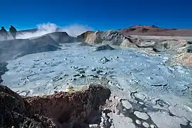 A pool of greyish-blue bubbling sludge surrounded by rocks and steam