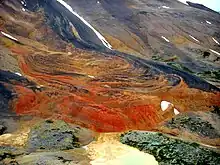 Orange, red and dark-colored rocks exposed on the side of a mountain.