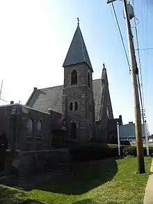 A brown sandstone tower stands in front of a church with a sharply pointed roof.