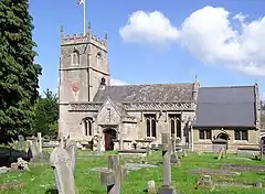 Yellow stone building, with porch with triangular roof in front. Short square tower with battlements topped by flag and flag pole. Gray gravestones in the foreground.