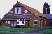 Brick-faced church with a 45-degree tiled gable roof, with a large front-mounted cross on a white background, on grassy land, c. 2004.