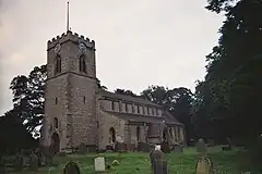 A small church with a square Ashlar tower, in the Early English style. The tower is to the left, the nave to the right, and we are looking somewhat obliquely at it.  The church is ringed by medium size trees, and a large Yew dominates the right of the picture, in the middle distance.  A handful of mismatched gravestones dot the grassy nearground of the picture.