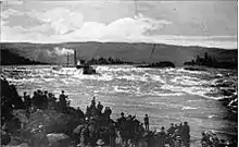 A river boat with more than a dozen windows along its visible side runs a set of rapids on a very large river. Smoke or steam rises from its smokestack and flows behind the boat parallel to the water. In the foreground, a crowd of 50 people watch the boat from the rocky shore.