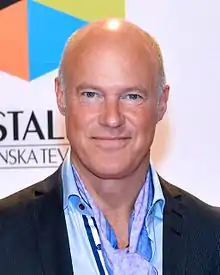 bald, middle-aged white male wearing a light-coloured shirt, dark suit jacket, and a neckerchief, looking directly at camera