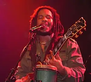 A man behind a microphone on a stand with his eyes closed, wearing a green jacket and holding a guitar.