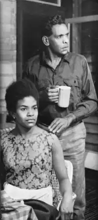 A black and white photograph of a male Aboriginal actor (standing) and a female Indigenous actor (seated) on the stage during the performance of a play