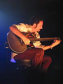 Lukather stands on stage, playing a black Ovation Adamas acoustic–electric guitar and singing into a microphone.