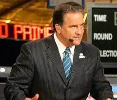 Candid photograph of Mariucci seated behind a desk on a TV set wearing a dark pin-striped suit, blue striped tie and a headset