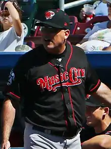 A man in a black baseball jersey with red trim and "Music City" written in red letters across the chest, a black cap with an "MC" on the front, and gray pants stands in front of a dugout.