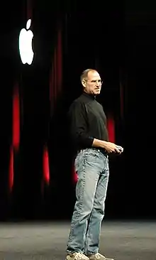 Full-length portrait of man about 50 years old wearing jeans and a black turtleneck shirt, standing in front of a dark curtain with a white Apple logo