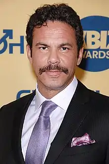 Portrait of a mustachioed man wearing a black coat over a white collared shirt and a silver patterned tie.