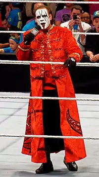 Sting wearing a red longcoat standing in a wrestling ring talking on a microphone