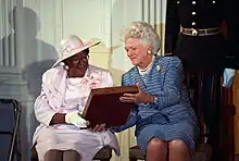 Bush, seated, hands a plaque to the woman sitting next to her.